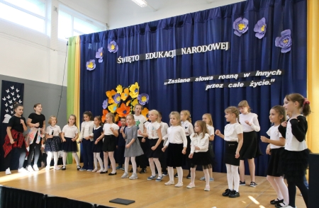 Uczniowie w strojach galowych, stoją w półkolu na scenie. W tle napis Dzień Edukacji Narodowej.