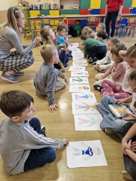 Dzieci w 2 rzędach siedzą naprzeciw siebie na podłodze i patrzą na wykonane rysunki. Obok pochyla sią nauczycielka.
