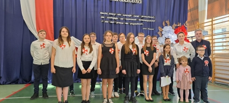 Uczniowie w strojach galowych na tle dekoracji. W tle flaga biało - czerwona